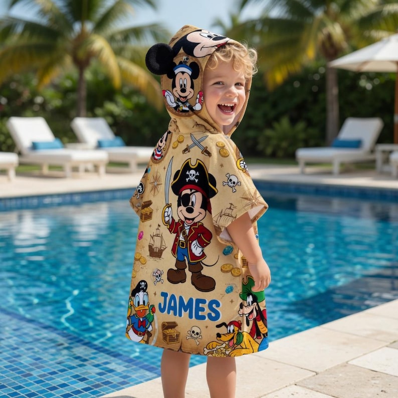 May include: A tan hooded beach towel featuring a pirate-themed Mickey Mouse design. The towel has a hood with Mickey Mouse ears and a pirate Mickey Mouse graphic on the front. The name "JAMES" is printed on the towel, along with other Disney characters.