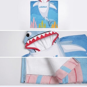 May include: A blue shark-themed hooded towel with a shark face hood and a shark body design. The towel features the name "JAMES" in blue letters. The towel has a white interior and is trimmed with pink and blue stripes.