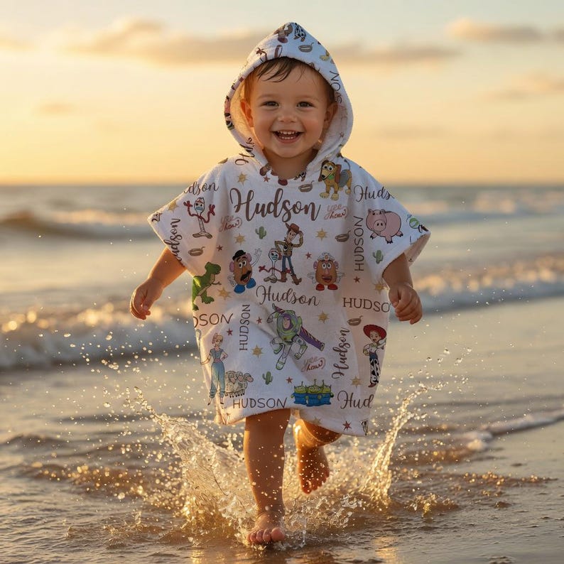 May include: A white hooded poncho towel featuring a repeating pattern of Toy Story characters including Woody, Buzz Lightyear, and Mr. Potato Head, interspersed with the name Hudson in a brown script font and small gold star accents.