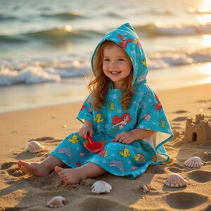 May include: A turquoise hooded beach towel featuring a colorful print of Ariel from The Little Mermaid, dolphins, and other sea creatures. The towel is being worn by a young child on a sandy beach.