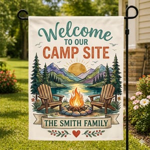 以下が含まれることがあります： 「Welcome to our Camp Site」の文字が書かれたガーデンフラッグ。水彩画で描かれたキャンプファイヤー、湖、山、2つの木製椅子が描かれています。バナーには「The Smith Family」と書かれています。
