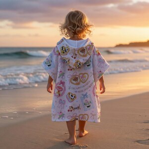 May include: A child wearing a white hooded beach towel with a colorful pattern of cartoon images, bows, and hearts, walking on a sandy beach. The towel has a hood and is decorated with pink, blue, and yellow accents. The background shows the ocean and a sunset.