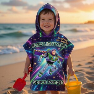May include: A child wearing a purple and blue Buzz Lightyear hooded beach towel with the text "Buzz Lightyear" and "Space Ranger". The child is holding a red spade and a yellow bucket on a sandy beach.