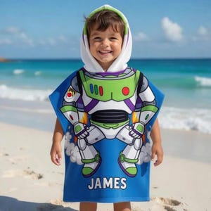 May include: A blue hooded beach towel featuring a Buzz Lightyear design. The towel has a white hood with green trim and the name "JAMES" printed in white. The background shows a beach and ocean.