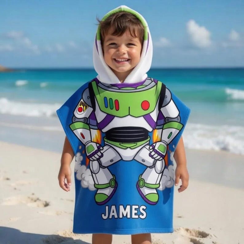 May include: A blue hooded beach towel featuring a Buzz Lightyear design. The towel has a white hood with green trim and the name "JAMES" printed in white. The background shows a beach and ocean.