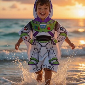May include: A white hooded beach towel with a Buzz Lightyear costume design, featuring green, purple, and white details. The towel displays a graphic of the character's spacesuit and the name "JAMES". The child is in the water, with the sea and sunset in the background.