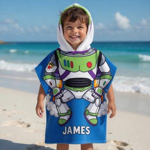 May include: A blue hooded beach towel featuring a Buzz Lightyear design. The towel has a white hood with green trim and the name "JAMES" printed in white. The design includes green, white, and purple accents, with a rocket ship graphic.