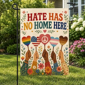 Op de afbeelding: Een tuinvaandel met de tekst "HATE HAS NO HOME HERE" in kleurrijke letters. De vlag toont handen die hartvormige borden vasthouden met verschillende ontwerpen, waaronder een regenboog, de Amerikaanse vlag, een vredessymbool en een transgender vlag.