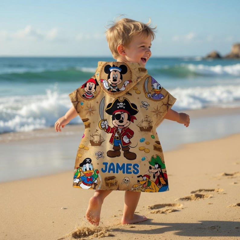 May include: A tan hooded beach towel featuring Disney characters in a pirate theme. The towel has images of Mickey Mouse, Donald Duck, and Goofy. The name "JAMES" is printed on the front. The child is on a sandy beach.