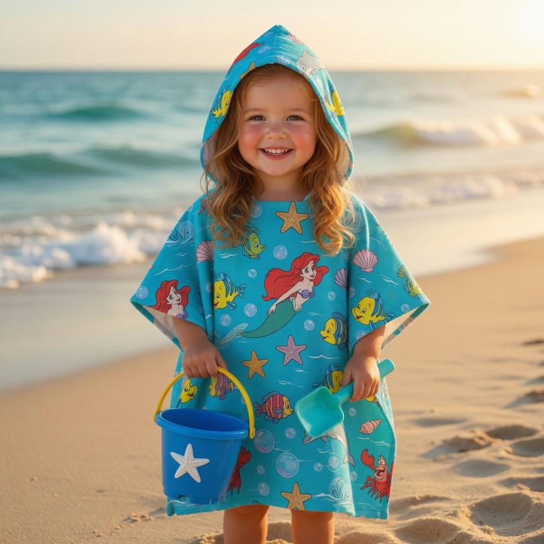 May include: A turquoise hooded beach poncho featuring Ariel from The Little Mermaid, starfish, and other sea creatures. The child is holding a blue bucket and shovel on a sandy beach. The poncho is made of soft, absorbent material.