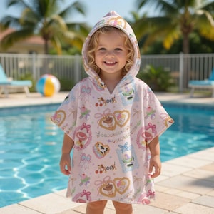 May include: A light pink hooded poncho with a colorful pattern of cartoon characters, bows, and hearts. The poncho is worn by a young child standing near a swimming pool. The child is smiling and looking at the camera.