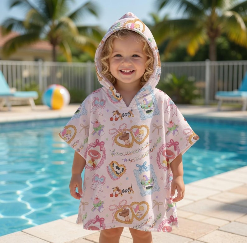 May include: A light pink hooded poncho with a colorful pattern of cartoon characters, bows, and hearts. The poncho is worn by a young child standing near a swimming pool. The child is smiling and looking at the camera.