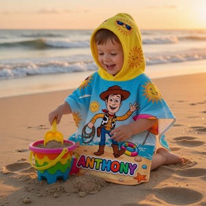 May include: A child wearing a blue and yellow hooded beach towel with a cartoon cowboy design, playing with a colorful bucket and shovel on a sandy beach. The towel features the name "ANTHONY". The hood has a sun design.