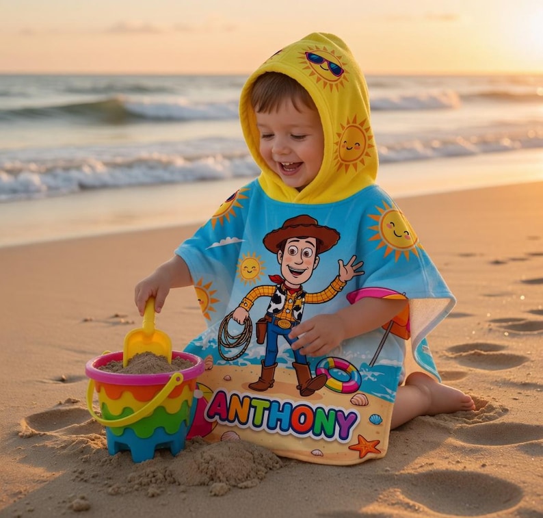 May include: A child wearing a blue and yellow hooded beach towel with a cartoon cowboy design, playing with a colorful bucket and shovel on a sandy beach. The towel features the name "ANTHONY". The hood has a sun design.