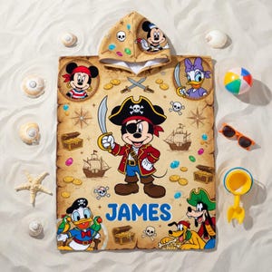 May include: A beige pirate-themed hooded towel featuring Mickey Mouse, Minnie Mouse, Donald Duck, Daisy Duck, and Goofy. The towel has a treasure map design with the name "JAMES" printed in blue. Additional beach toys and seashells are scattered around the towel.