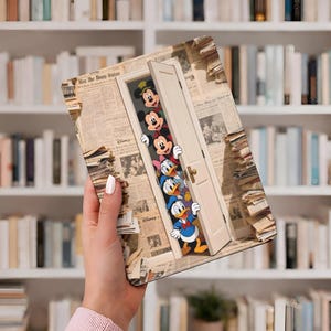 Może przedstawiać: Prostokątne etui na tablet z fantazyjnym wzorem przedstawiającym postacie Disneya wyglądające zza białych drzwi. Etui ma tło w stylu gazety i zawiera Myszkę Miki, Minnie, Kaczora Donalda i Daisy Duck.