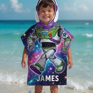 May include: A hooded beach towel featuring a colorful graphic of Buzz Lightyear from Toy Story, with a space-themed background. The towel has a white hood with purple trim and the name "JAMES" printed on the bottom.