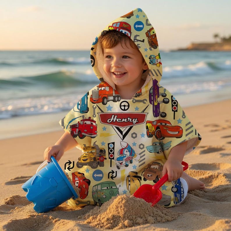 May include: A child wearing a yellow hooded beach towel with a colourful car and truck pattern. The towel has the name "Henry" on it. The child is playing with a blue bucket and a red shovel on a sandy beach.