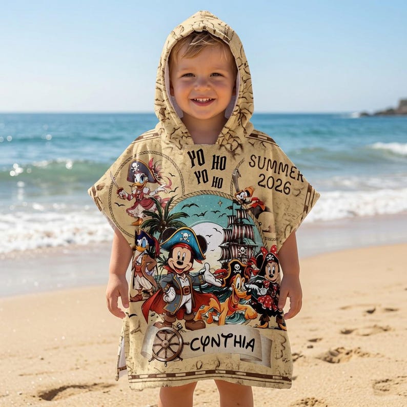 Op de afbeelding: Een beige poncho met capuchon voorzien van een piratenthema met Mickey Mouse, Minnie Mouse, Donald Duck en Pluto. Het ontwerp bevat een schip, palmbomen, de tekst "YO HO YO HO", "SUMMER 2026" en de naam "CYNTHIA" op een perkamentrol.