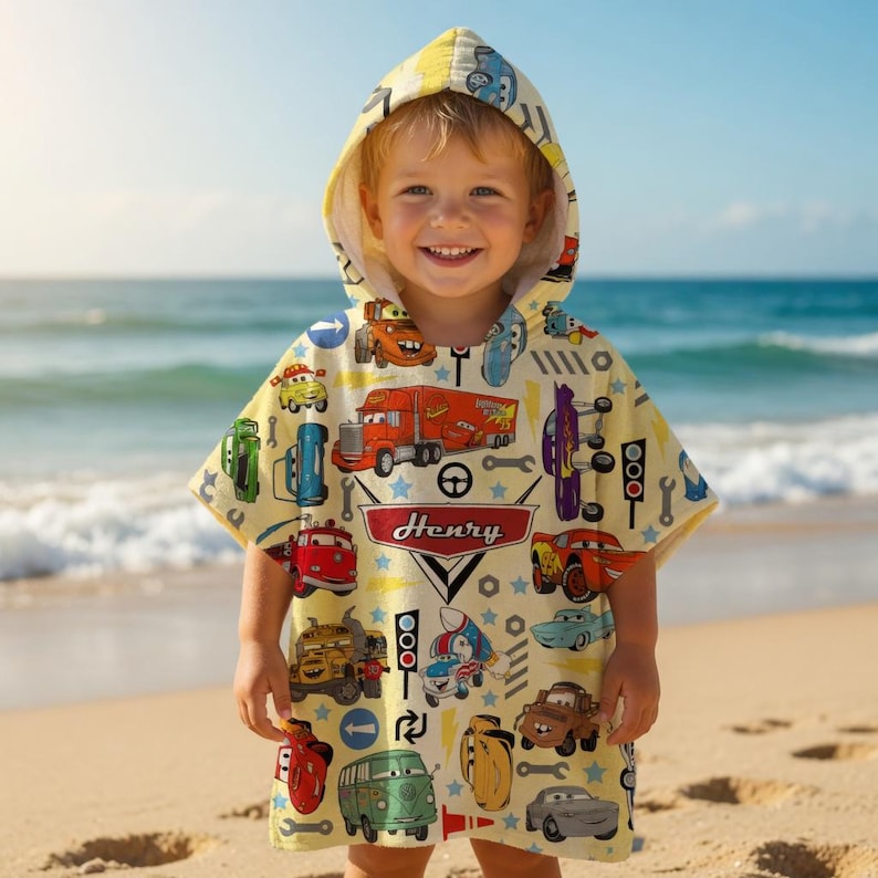 May include: A child wearing a yellow hooded beach towel with a colourful car design. The towel features various cartoon cars, trucks, and tools. The name "Henry" is printed on the towel. The child is smiling on a sandy beach with the ocean in the background.