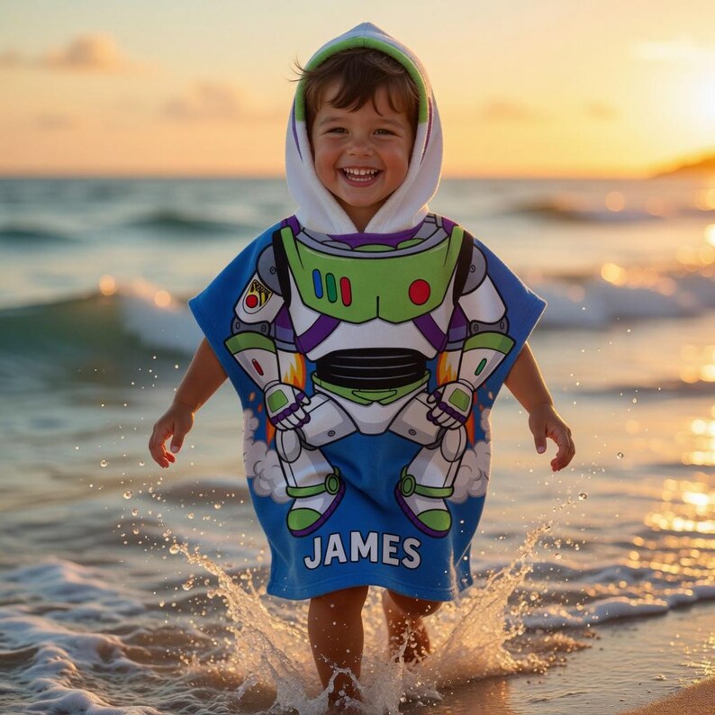 May include: A blue hooded beach towel featuring a Buzz Lightyear design. The towel has a white hood with green trim and the name "JAMES" printed on the bottom. The child is wearing the towel while playing in the ocean.