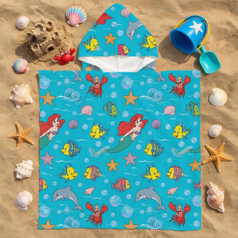 May include: A hooded beach towel with a vibrant turquoise background featuring Ariel, Flounder, Sebastian, and other characters from The Little Mermaid. The towel is surrounded by seashells, starfish, and a blue bucket with a shovel on a sandy beach.
