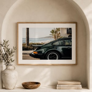 May include: A framed print featuring a dark green vintage sports car parked on a street with a coastal backdrop. The car has a rear spoiler and black wheels. The print is displayed on a shelf with a vase, wooden bowl, and books.