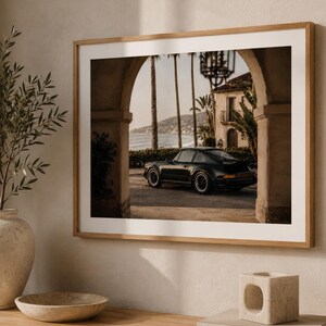 May include: A framed wall art print featuring a dark-coloured sports car parked under a stone archway. The scene includes tall palm trees, a coastal view with the ocean in the background, and a Mediterranean-style building with a balcony.