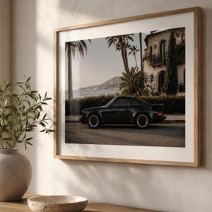 May include: A framed wall art print featuring a side profile of a black sports car parked on a street. The background shows a coastal scene with palm trees, a white building with arched windows, and a distant mountain under a soft sky.