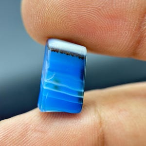 Puede incluir: Una pequeña piedra preciosa pulida de forma rectangular con bandas horizontales de color azul vibrante y blanco. La piedra muestra una superficie lisa y brillante con una línea oscura y moteada que separa la sección blanca superior.