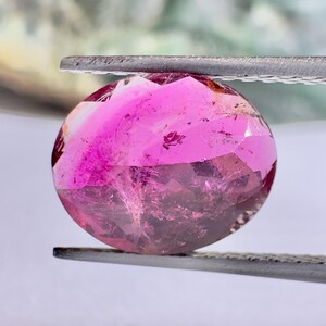 May include: A faceted, round gemstone held by tweezers. The gem is a vibrant pink color with darker, purplish inclusions. The light catches the facets, creating a sparkling effect. The tweezers are silver.