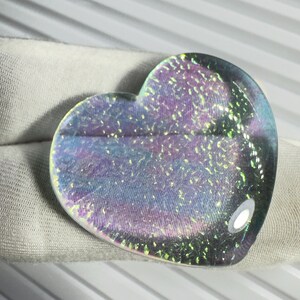 May include: A heart-shaped, iridescent glass object with a shimmering, multi-coloured surface. The colours include shades of purple, blue, and green. The object is resting on a white fabric surface.
