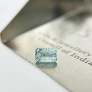 Op de afbeelding: Een rechthoekige, lichtblauwe, gefacetteerde edelsteen rustend op een metalen oppervlak. Het oppervlak bevat gedeeltelijk zichtbare gegraveerde tekst die luidt Gem & Jewellery Council of India. De steen is doorschijnend met zichtbare insluitingen.