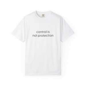 Puede incluir: Una camiseta blanca de manga corta con el texto negro "control is not protection" impreso en una fuente sans-serif sencilla en el centro del pecho. La prenda tiene cuello redondo y un corte estándar con una etiqueta de marca visible en el cuello.