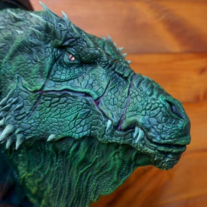 以下が含まれることがあります： 詳細なドラゴンの頭の彫刻。主に緑色で、質感のある鱗と鋭い白い歯があります。ドラゴンは、見える赤い目をして、恐ろしい表情をしています。彫刻は木製の背景にあります。