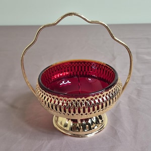 Può includere: Una ciotola in vetro rosso rubino inserita in una cornice in metallo dorato con filigrana. La ciotola presenta una base circolare e un manico alto e arcuato dal design smerlato. La struttura metallica circonda l'esterno del vetro, creando un cestino.