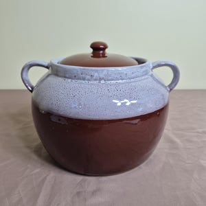 May include: A round ceramic pot with two side handles and a matching lid. The lower portion of the pot and the lid are a solid dark brown, while the upper portion features a textured, speckled light gray glaze that transitions into the brown.