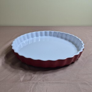 May include: A round ceramic tart dish featuring a glossy white interior and a matte red exterior. The bakeware has a classic fluted edge design, providing a scalloped rim that creates a decorative border for baked goods and various culinary preparations.