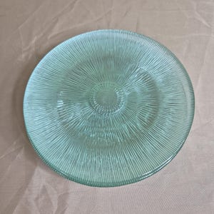 May include: A round, light green glass plate featuring a textured surface with a radiating sunburst pattern of fine, raised lines extending from a smooth circular centre to the outer edge of the rim, creating a distinct light-refracting effect.