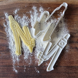 Può includere: Due stampini per biscotti in plastica bianca a forma di compasso da disegno, uno dei quali ha impresso una forma di pasta giallo pallido nella farina, mentre l'altro rimane vuoto, mostrando il contorno dettagliato dello strumento da disegno.