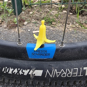 Puede incluir: Un tapón de válvula amarillo brillante con forma de plátano fijado a la válvula de un neumático de bicicleta. El tapón se sitúa sobre una base rectangular azul con el texto PRESTA SCHRADER COMPATIBLE impreso en negro sobre el neumático.
