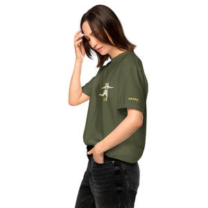 Könnte beinhalten: Olivgrünes T-Shirt mit einem Fußballspieler-Motiv auf der Vorderseite und dem Wort "HEAPS" auf dem Ärmel. Die Grafik zeigt einen Fußballspieler beim Schießen eines Balls. Das T-Shirt ist aus weichem Material.
