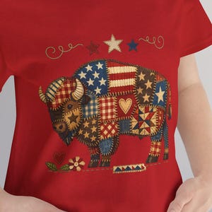 Op de afbeelding: Rood T-shirt met een patchwork buffelontwerp met sterren, harten en Amerikaanse vlagmotieven. De buffel is versierd met diverse patronen en kleuren, waaronder blauw, rood en bruin. Het shirt heeft een ronde hals en korte mouwen.