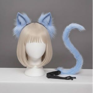 Op de afbeelding: Een lichtblauwe kattenoren-hoofdband en staartset. De oren zijn pluizig en bevestigd aan een zwarte hoofdband. De staart is lang, gebogen en heeft een zwarte verstelbare riem met een gesp. De set wordt getoond op een mannequinhoofd met een blonde pruik.