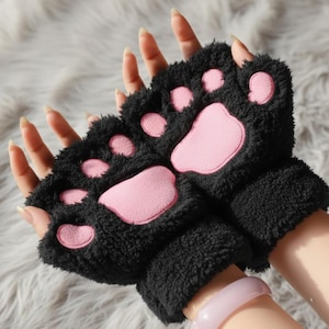Peut inclure: Gants sans doigts noirs avec des coussinets roses en forme de pattes. Les gants sont faits d'une matière douce et duveteuse. Les coussinets sont de couleur rose clair et cousus sur les gants noirs. Les gants sont conçus pour ressembler à des pattes de chat.