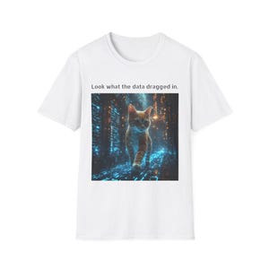 Puede incluir: Camiseta blanca con una impresión de arte digital. La impresión muestra un gato atigrado naranja caminando en una escena de ciudad futurista. El texto sobre la imagen dice: "Look what the data dragged in."