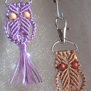 May include: Two macrame owl keychains. One is lavender with wooden bead eyes and a matching tassel. The other is tan with brown bead eyes and a tan tassel. Both have silver key rings and clasps.