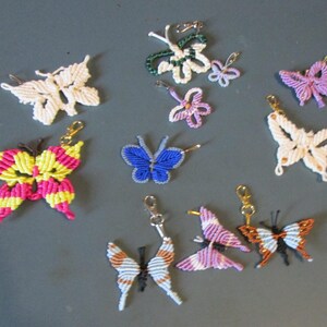 May include: A collection of handmade macrame butterfly accessories in various colors. Includes keychains, pins, and decorative pieces. Colors include white, pink, yellow, blue, purple, orange, and black. The butterflies have intricate knotting patterns.