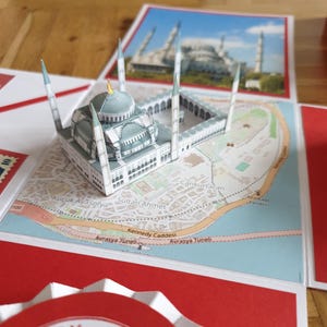 Könnte beinhalten: Ein Papiermodell der Blauen Moschee in Istanbul, Türkei, liegt auf einer Karte. Das Modell ist hellblau und weiß, mit Minaretten und einer Kuppel. Die Karte zeigt die Umgebung mit sichtbaren Straßennamen.