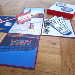 Könnte beinhalten: Eine offene Geschenkbox mit ABBA Voyage Branding, inklusive eines Londoner Busses und einer Telefonzelle aus Papier auf einer Karte, zwei gedruckten Tickets mit der Aufschrift YOU ARE GOING TO ABBA Voyage und einem quadratischen Deckel.